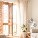 Les 7 meilleures astuces sans clim pour la santé et l’habitat pendant la canicule en 2026 Intérieur lumineux d’un appartement en période de forte chaleur, avec volets fermés, rideaux clairs et ventilateur discret dans une ambiance estivale réaliste.